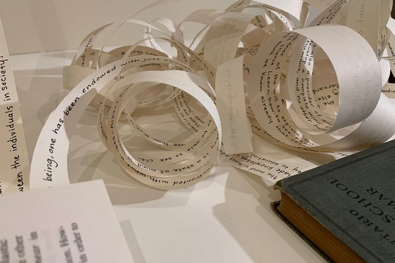 Art installation of three books lying on the floor with strips of paper coming from inside the books and stretched up the wall