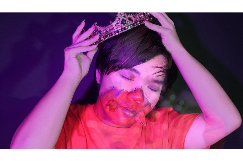 Person, lit by a pink light and with red paint on the lower half of their face, positions a crown on their head.