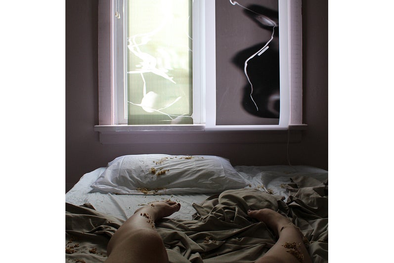 Mattress, rumpled sheets and a person's feet on the floor in front of window panes covered with screen with smoke-like pattern