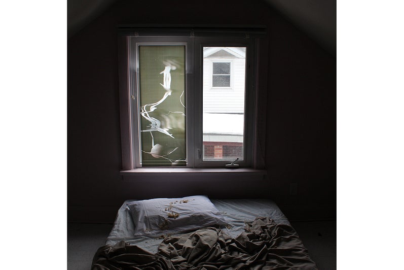 Dark room with a mattress and rumpled sheets on the floor in front of a window.  Window panes are covered with translucent film