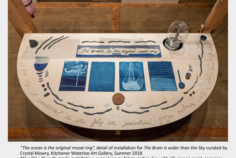 Art installation on table with various objects and 4 pages in shades of blue showing mermaid, stars, boat and waves. Words at to