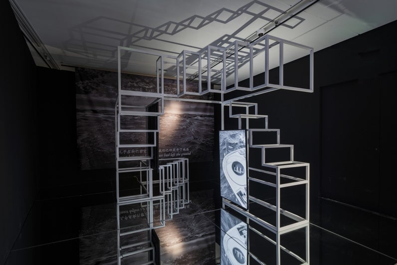 Art work in a dark room with a projection on the back wall of grey scale story sea with text at the bottom.  In the center of the room is a scaffold stair-like structure.