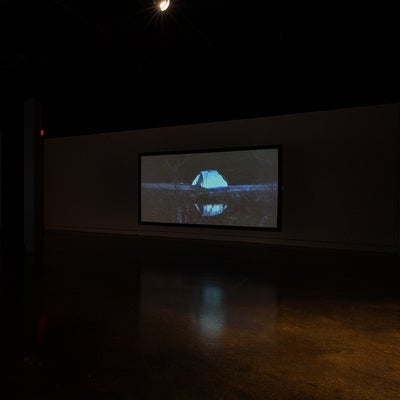 Video exhibition in dark gallery.  Video on left shows hands carving letter "D", back wall video of glowing blue tent by water.