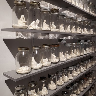 Six shelves filled with mason jars containing sculptures of white seated figures.
