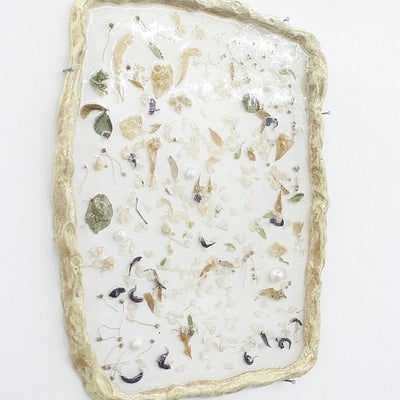 Flat, rectangular ceramic artwork hung on a wall.  Work is glossy white with flower petals, leaves and pearl embedded in surface