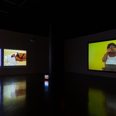 Video exhibition in dark gallery. Videos at left show person lying on the floor with head covered in rice and white cotton balls