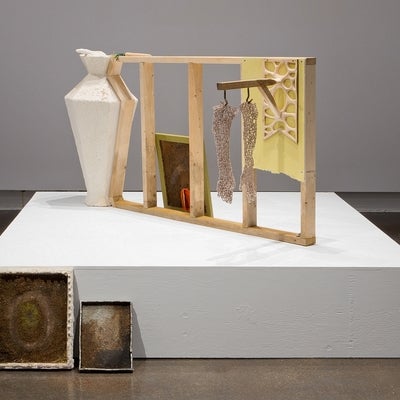 Art installation of various objects on and around a white platform.  Objects include several earthtone paintings, plaster vase, 