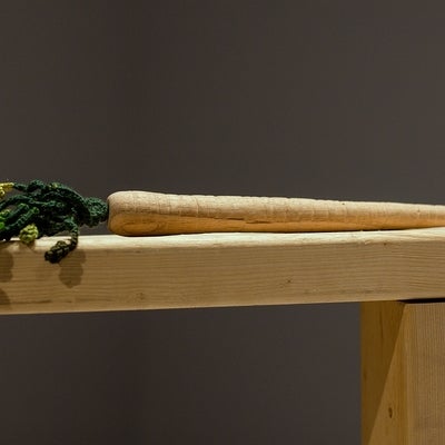 Artwork showing a wooden carrot with cotton crocheted leaves sits on a wooden board.
