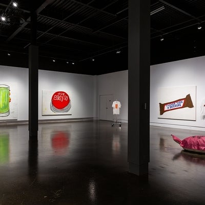 Exhibit with 3 large colourful paintings of a glass of green juice, red button and a chocolate bar, t-shirts and floor cushion