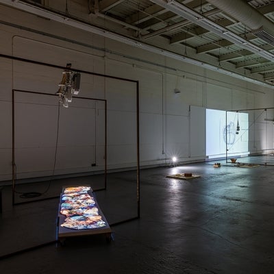 Dark, industrial space with art installation. Projectors light objects on the concrete floor and a video of a person drawing.