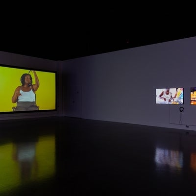  Video exhibition in dark gallery. Video at left show person in a chair cutting sections of their hair, at right videos show peo