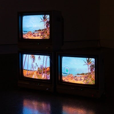 Video exhibition in dark gallery. Two small monitors on floor with one more stacked on top, all show videos of tropical landscap