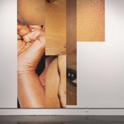 Art exhibition in gallery. Large scale photographs on walls depict details of a black woman's body. 