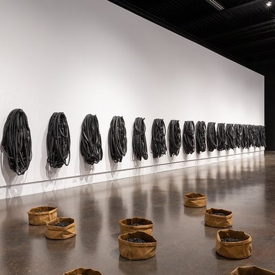 Artwork in a gallery. On walls, multiple groups of bike tubes. On floor, series of large round paperbags with asphalt