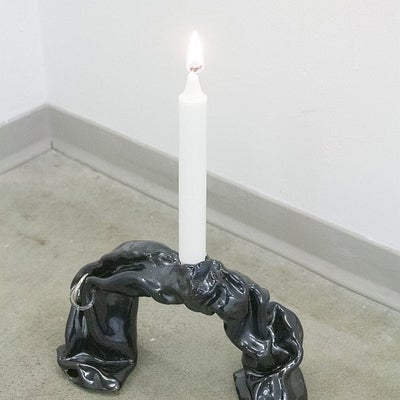 Ceramic sculpture resembling crumpled black plastic in the shape of an upside-down "u" with a lit, white candle on the top.