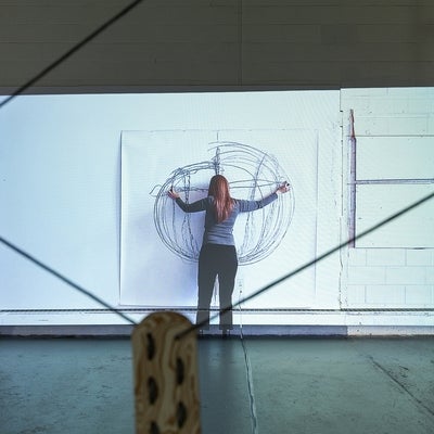 Video of a person drawing on large paper hung on a wall.  The person draws a circular shape using each arm in a sweeping motion,