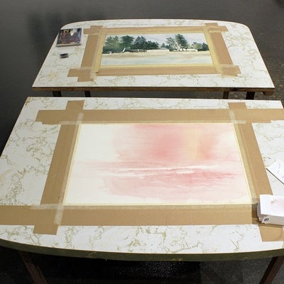 Two small formica-topped tables, seen from above, with a watercolour painting taped to each. Top painting of evergreen trees, 