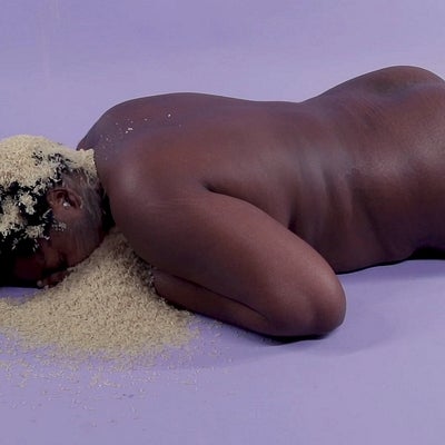 Video still of person lying nude, face down on the floor with rice covering their head