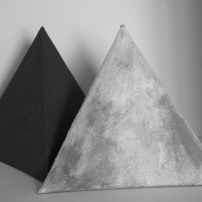 Two small pyramids with rough, irregular surfaces. The pyramid in front is shades of light grey, the one behind is black.