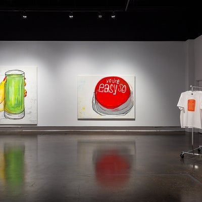 Exhibit of paintings of a glass of green juice and of a red button reading “you’re so easy”, and a rack of t-shirts with printed