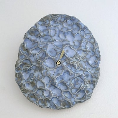 Flat, round, blue and grey ceramic plaque with a dimple surface and a single brass clock hand in the middle, hangs on a wall.