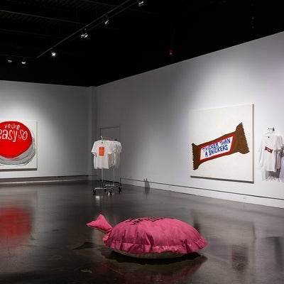 Art exhibit with 2 large paintings, t-shirts and a large pink cushion on the floor.