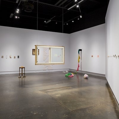 Art gallery installation of multiple ephemera, white ceramic tiles, shells, crocheted items and a salvaged house window hanging in the center of the room.