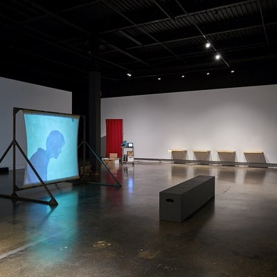 Art gallery installation with videos, one projected on a free-standing screen, wooden drawers attached side by side on a wall and a metal cart and privacy screen in the corner.