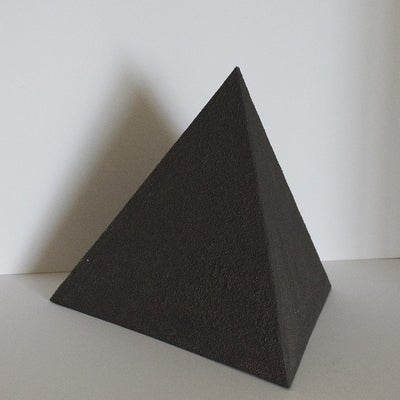 Small, black. pyramid with rough, irregular surfaces in front of a white wall.