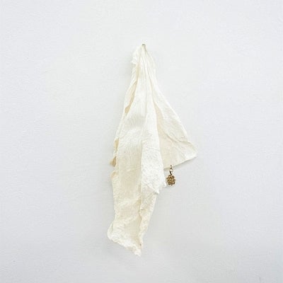 Handmade paper resembling a white cotton cloth, and a small gold pendant, hangs on a white wall.
