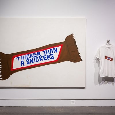 Art exhibit of a painted chocolate bar labelled "Thicker than a Snickers", and t-shirts printed with the same image.