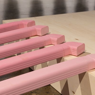 Detail of five cast pink sculptures resemble eavestrough downspouts sitting on the edge of a wooden riser on the gallery floor. 