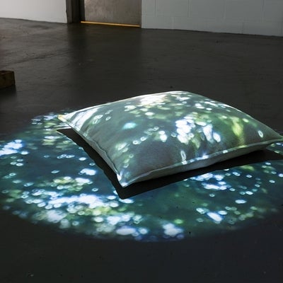 Artwork of a digital projector shining the image of dappled sunlight on a pillow on the floor.