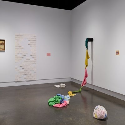 Art gallery installation of brightly coloured terry cloth towels sew together to form a rope, a crochet covered rock, white ceramic tiles, and a vintage landscape painting.