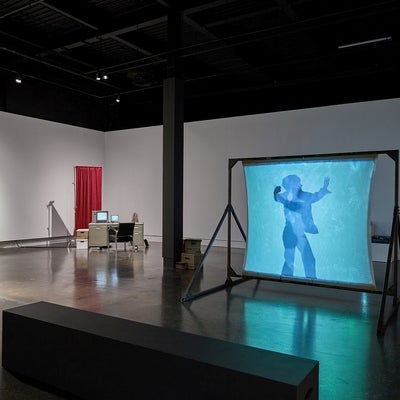 Art gallery with installation of videos, one projected on a free-standing screen, an industrial office desk, chair and privacy screen and multiple bankers boxes.