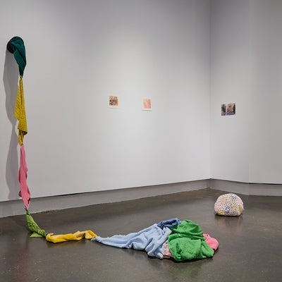 Art gallery installation of brightly coloured terry cloth towels sew together to form a rope, a crochet covered rock, and three small photo reproductions.