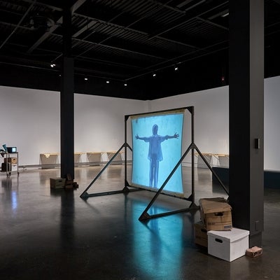 Art gallery installation with videos, one projected on a free-standing screen, wooden drawers attached side by side on a wall and a metal cart and privacy screen in the corner.