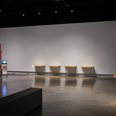 Art gallery with wooden drawers attached side by side on a wall and a metal cart holding a small television, boots and bankers boxes sits in front of a privacy screen.