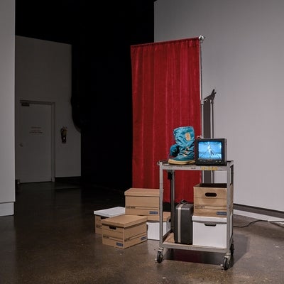 A metal cart with a small monitor, winter boots and bankers boxes, sits in front of a privacy curtain