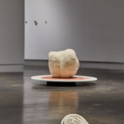 A large off-white porcelain peach pit rests on the floor in front of a spherical fabric sculpture on circular platform.