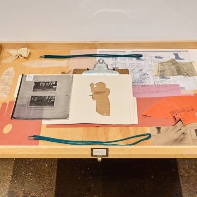 Wooden drawer displaying vintage plans, product catalogue pages, file folder, clipboard, and rubber condoms decorated with text.