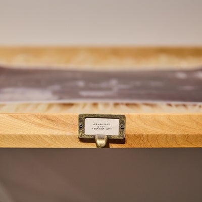 Detail of a wooden shelf with a brass drawer pull label reading "A.R. Kaufman is not a feminist icon".