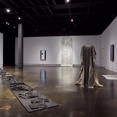 Art gallery exhibition with video monitors, large hanging drawings that drape onto the floor, a canvas dress hung from the ceiling and a row of blackened objects on the floor. 