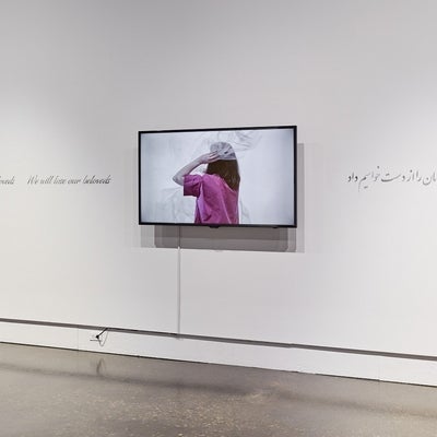 Art gallery exhibition with a video monitor and text in English (We will lose our beloveds) and Arabic running along the wall.