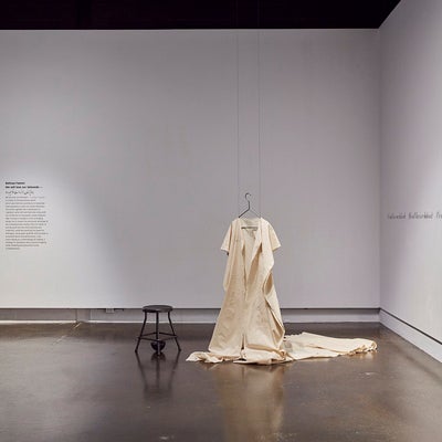 Art gallery exhibition text running along the wall, a canvas dress is hung from the ceiling with a blackened stool beside it.