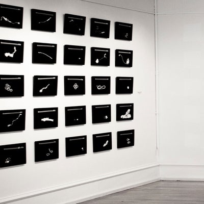 Gallery installation of artworks hung in a 5x5 grid.  Works are black and feature white shapes.