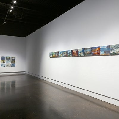 Exhibition of paintings showing two walls of a gallery. Wall on right has multiple small, colourful paintings hung in a line.  Paintings show various views of a pedestrian overpass between building. Wall on left shows twelve winter landscape paintings hung in a grid.