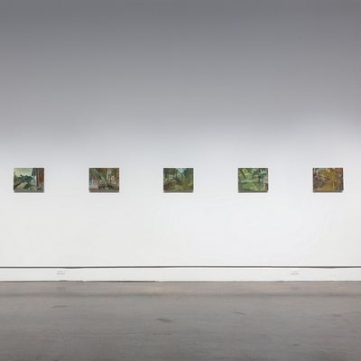 Exhibition of paintings in a gallery, showing seven small landscapes painting hung in a row.