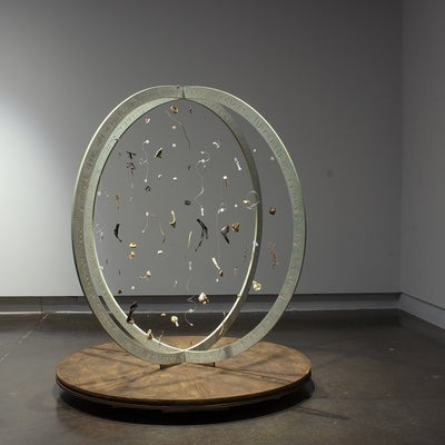 A sculpture of two intersecting circular structures, with words cut into the sides, sits upright on the floor with items hanging in the center of the sculpture.