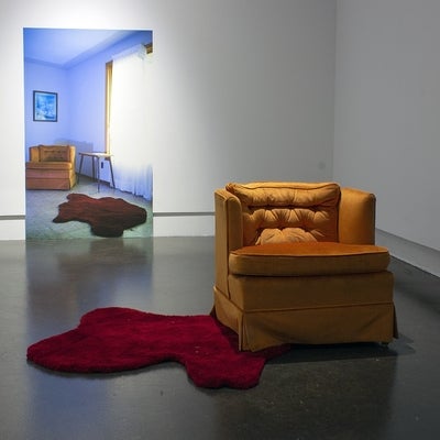 Art installation in a gallery with a large photograph of a living room interior with an orange armchair and red rug. In front is the actual orange armchair and an irregularly shaped red floor rug that resembles a pool of blood.